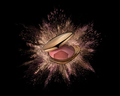 Un'esplosione di cipria colorata dietro la confezione d'orata del blush tricolore della Kiko. Sfondo nero. Orizzontale