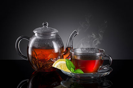 Una teiera di vetro con il tè caldo fiorito e a destra una tazza di vetro trasparente con la fetta di limone e foglia di menta sul piattino.